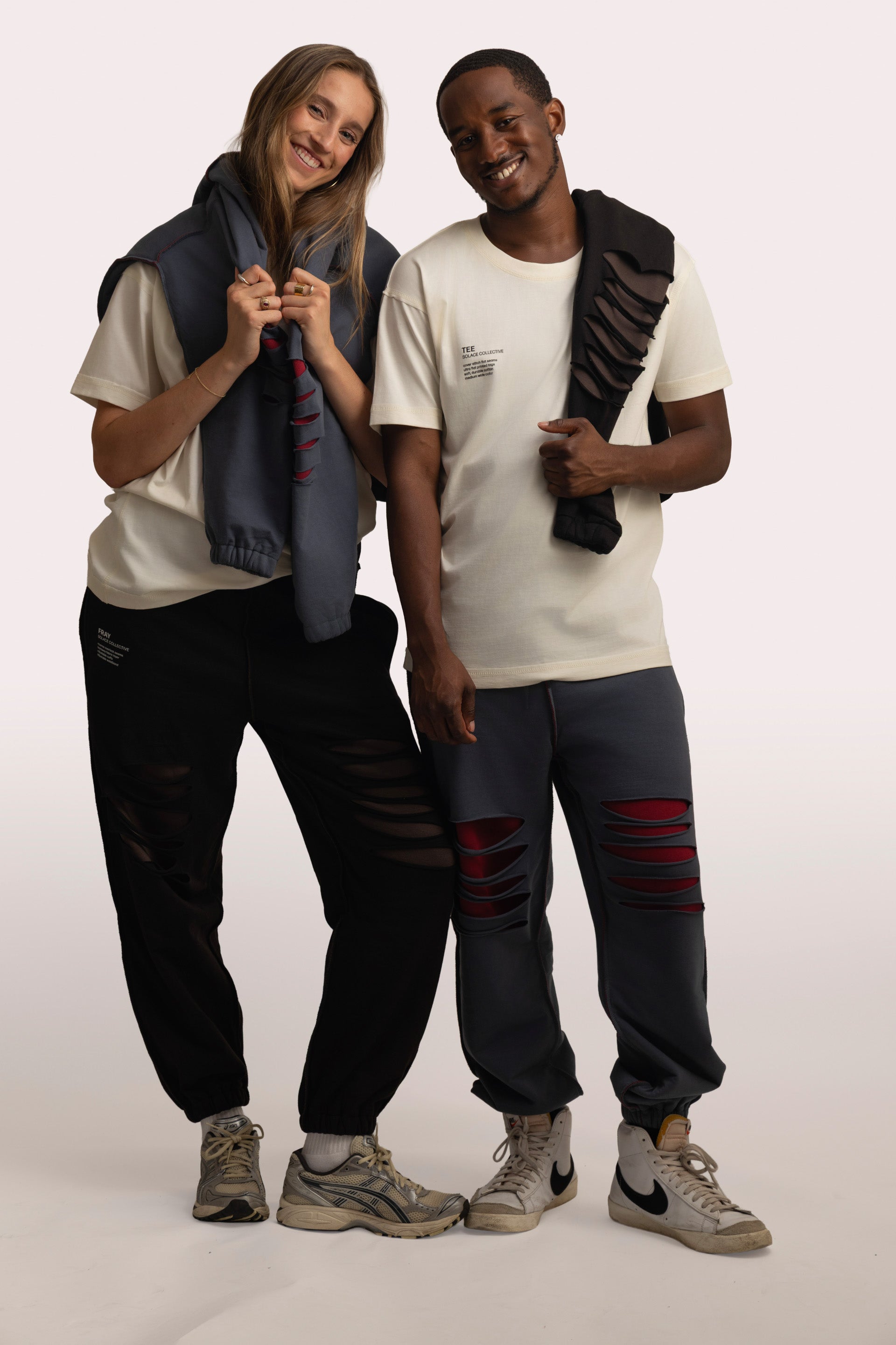 The image features a man and a woman wearing SOLACE Collective pieces styled in a casual, coordinated look. Both are dressed in ivory-colored SOLACE TEE t-shirts paired with the FRAY sets. Each has a SOLACE Collective FRAY hoodie draped over their shoulders, and they are standing closely together, smiling, showcasing the relaxed and expressive aesthetic of the collection.