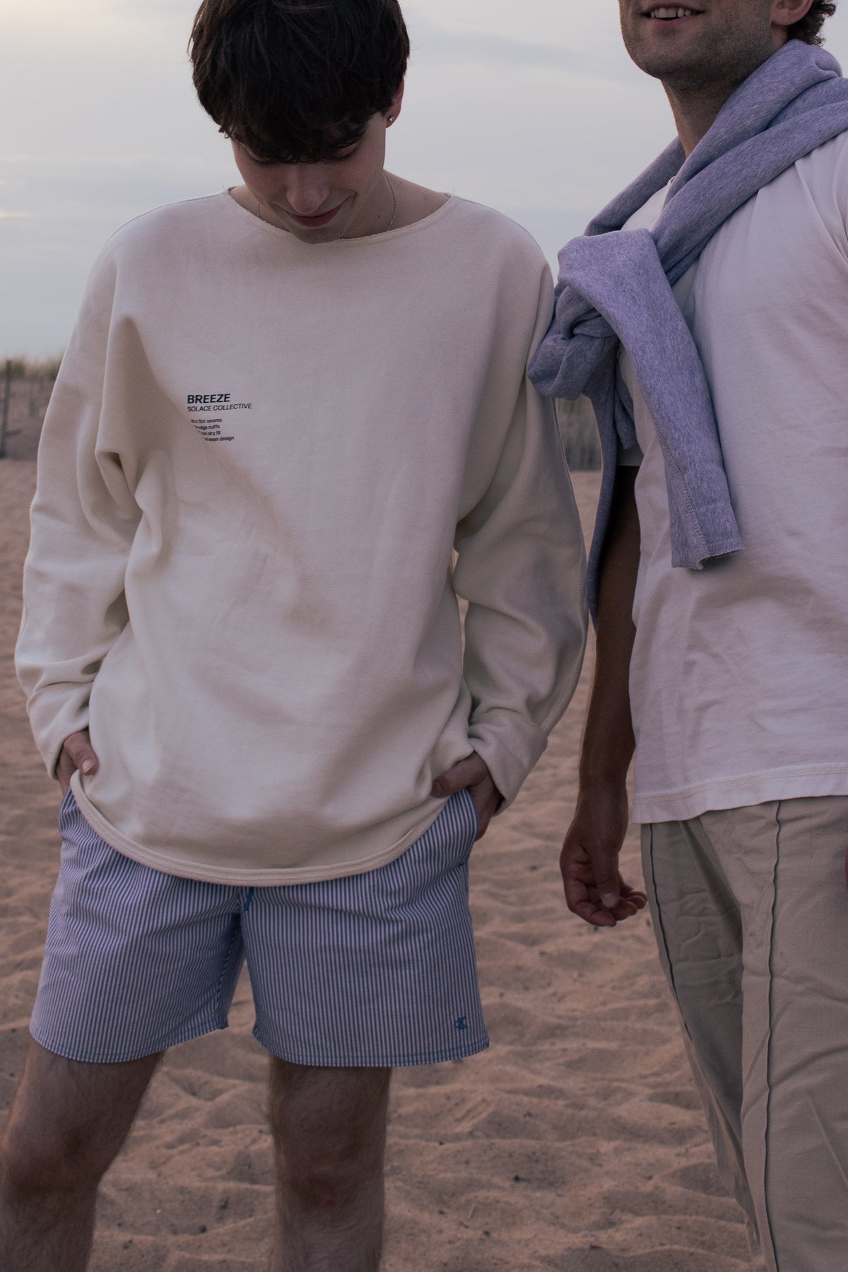 Two men standing on a sandy beach. The man on the left wears a cream-colored long-sleeve crewneck sweatshirt with a small black text graphic on the left chest and light blue pinstriped shorts.