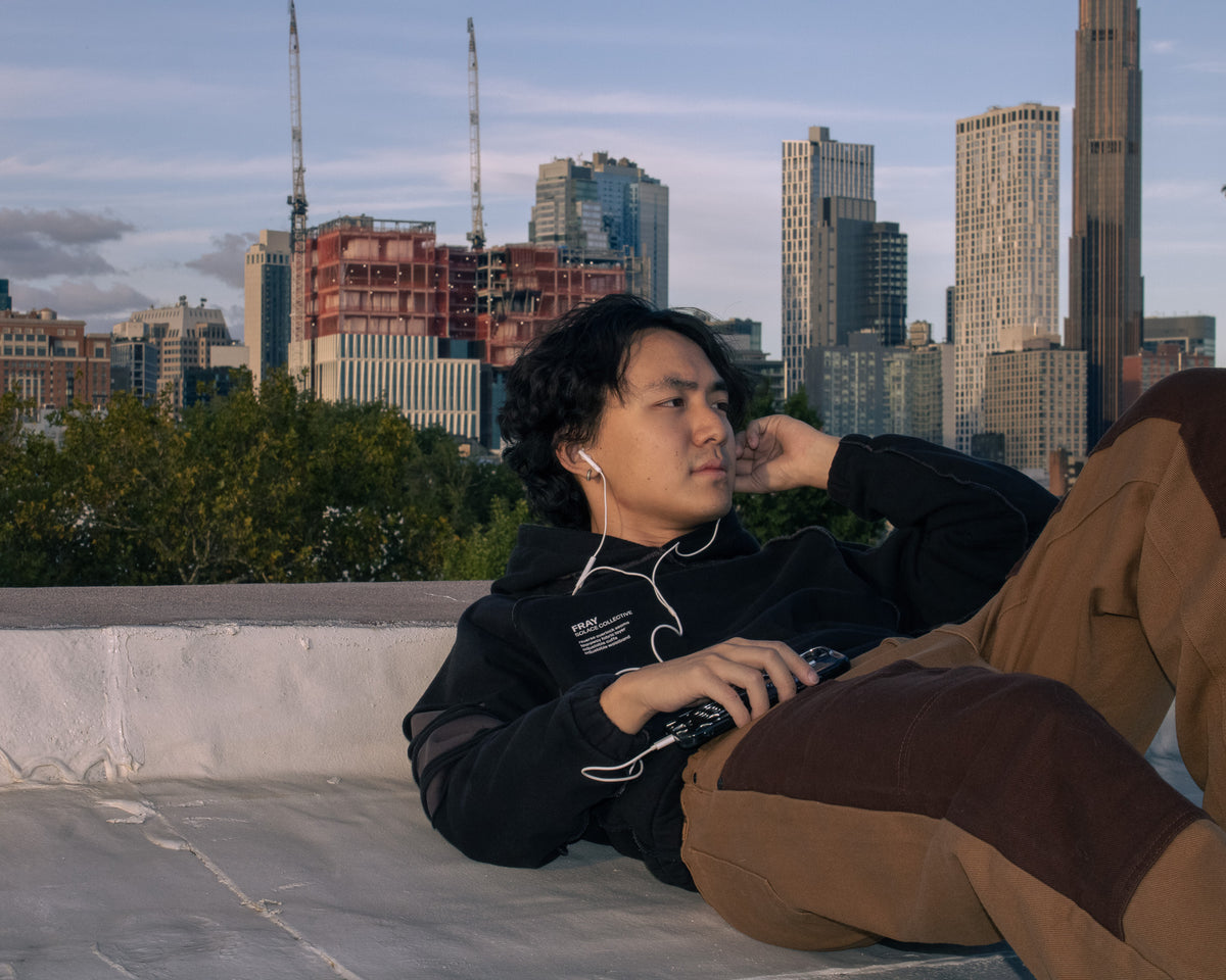 A man wearing a black hoodie and brown pants relaxes on a rooftop terrace with a city skyline in the background, wearing white wired earphones.